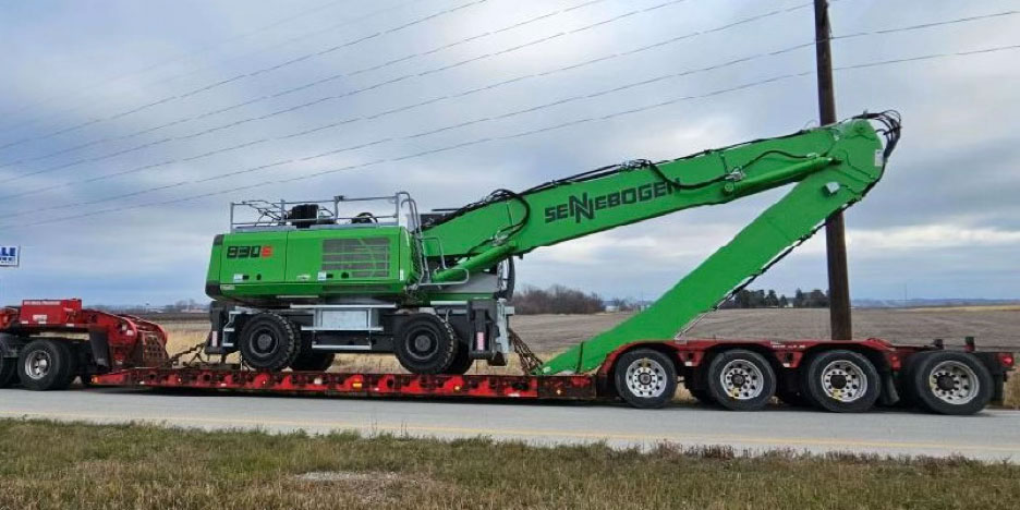 A side view of a green Sennebogen material handler loaded onto a flatbed trailer, parked on the side of a road against a backdrop of open fields.