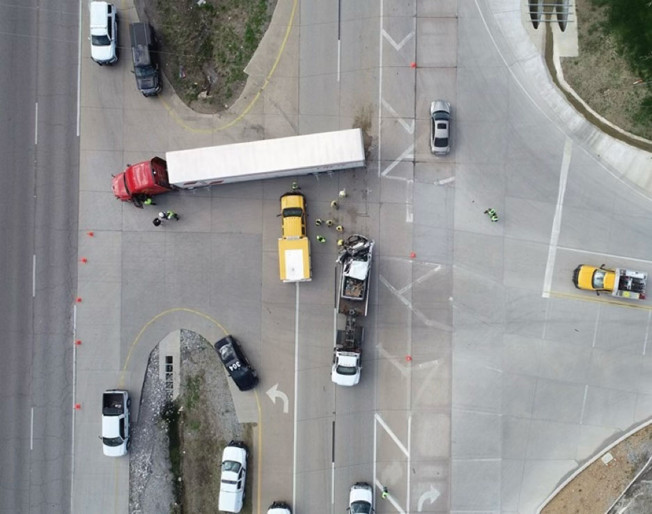 An aerial view of a traffic accident scene showing a red semi-truck, a stationary passenger vehicle, and emergency response personnel gathered on the pavement.