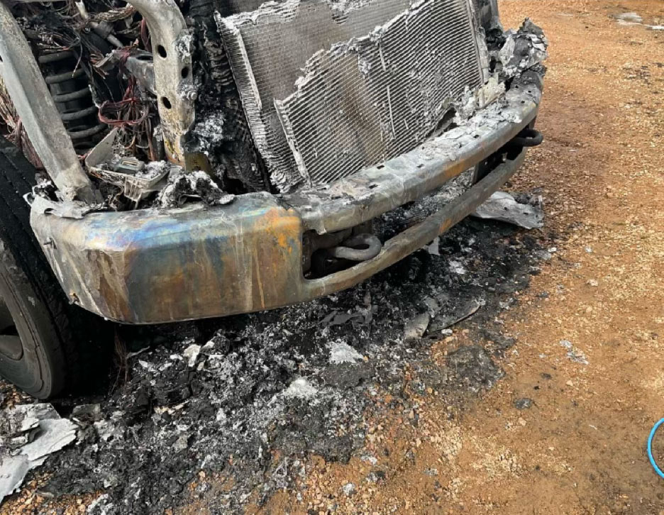 A view of the front end of a vehicle showing significant fire damage to the engine compartment, including a melted radiator and charred components, as part of a forensic investigation into a safety recall.