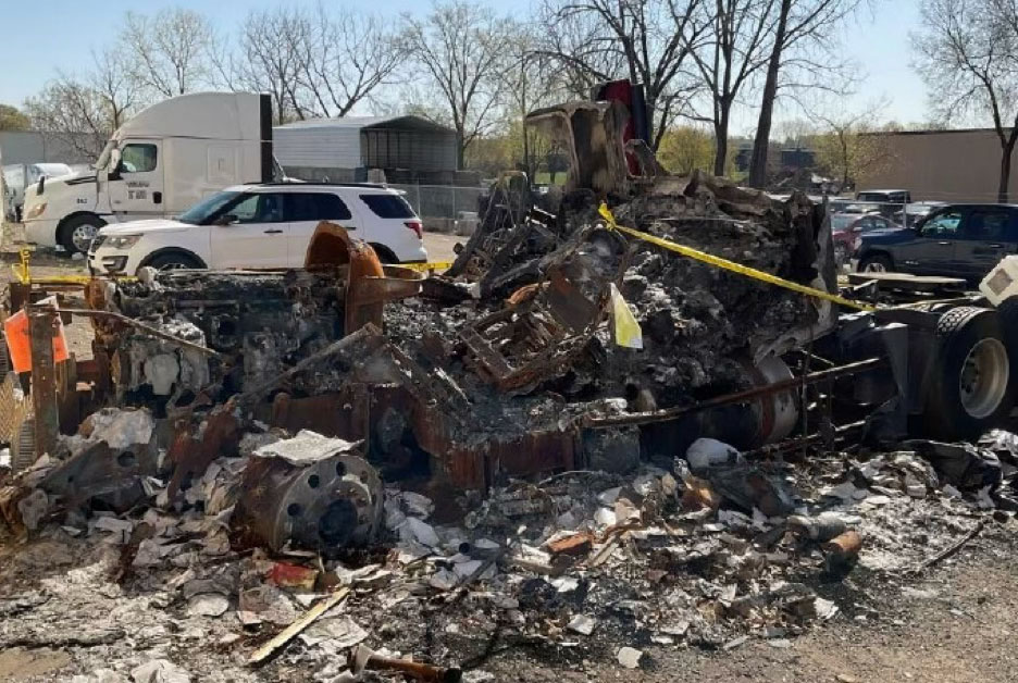 A charred, heavily damaged tractor-trailer unit remains at a scene, surrounded by yellow hazard tape, indicating the site of a vehicle fire investigation.