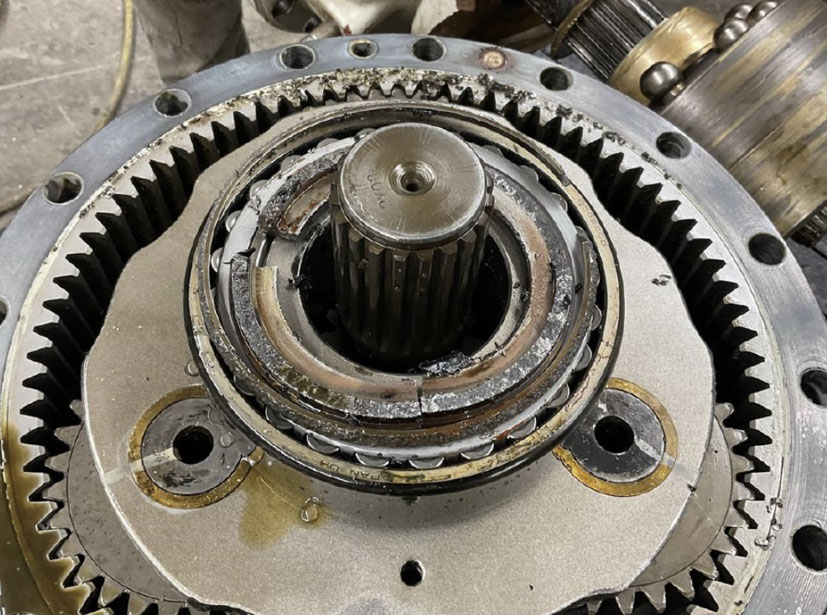 A close-up of a damaged planetary gear assembly showing significant wear, fractured teeth on the ring gear, and metal debris within the bearing housing, consistent with a catastrophic mechanical failure.